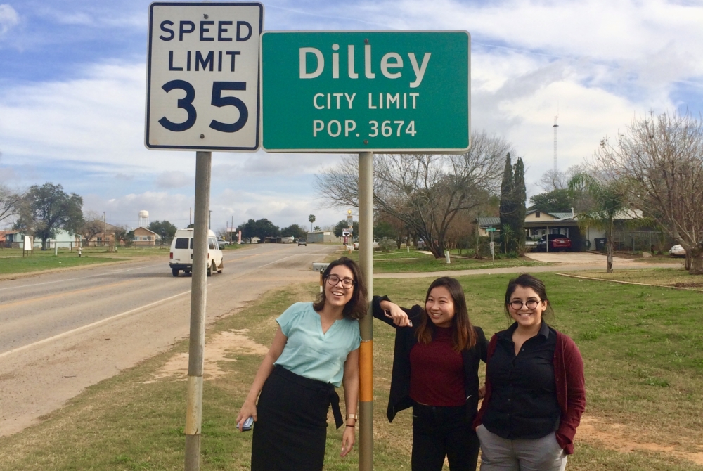 Three women stand by the side of a road in a rural town. The road sign above their heads reads “DILLEY CITY LIMIT: POP. 3674”