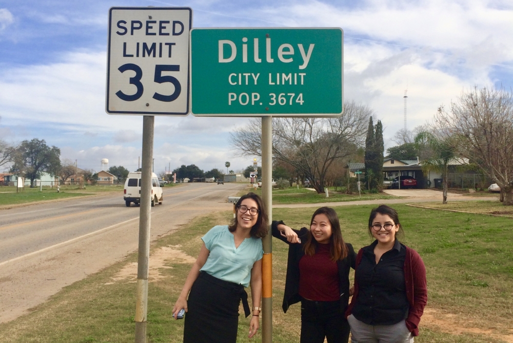 Three women stand by the side of a road in a rural town. The road sign above their heads reads “DILLEY CITY LIMIT: POP. 3674”